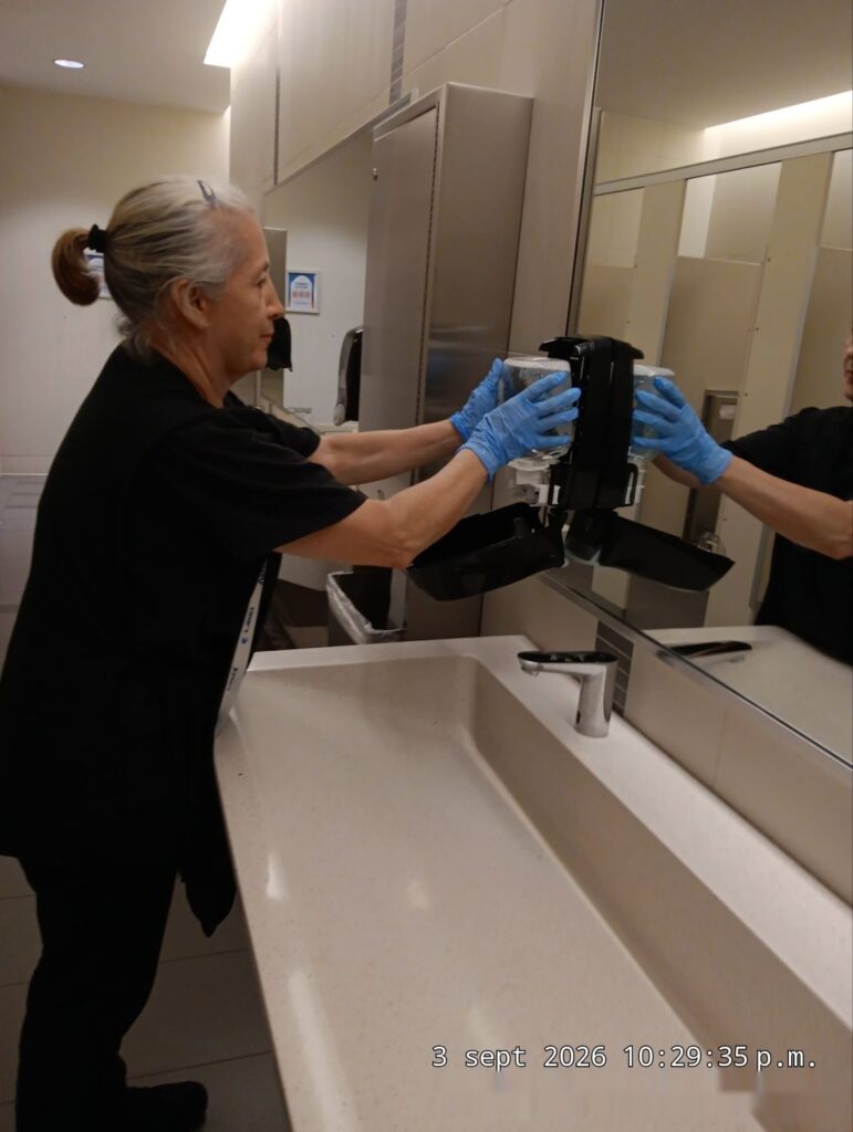 Caring Commercial Cleaning custodian wearing gloves refilling a touchless soap dispenser in a school restroom in Houston, TX